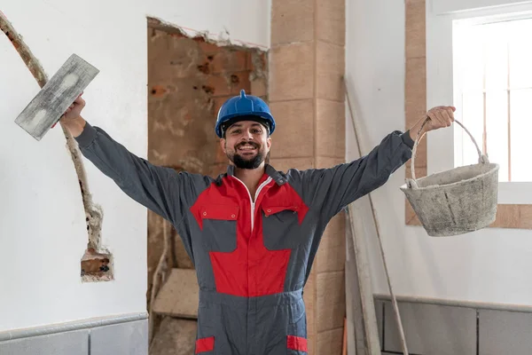 Kürekçi ve çimento sepeti ile tuğla ustası kolları havada kameraya bakıyor.