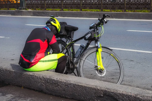 A cyclist rests on the edge of the road. A professional cyclist sits next to the bike. Bicycle breakdown on the road