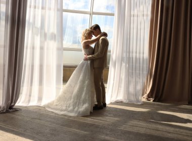 Yeni evliler büyük panoramik pencerenin arkasında duruyor. Romantik çift gelin ve damat. Gelin ellerini damadın omzuna koydu. 