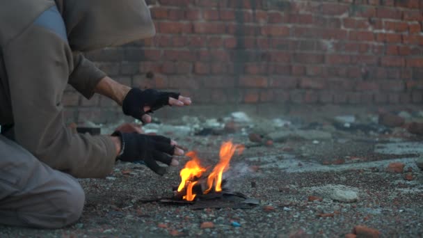 A homeless man warms his hands by the fire. Post-apocalypse scene. Hands in black torn gloves. Close-up. Weak warmth from a small fire — Stock Video