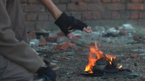 En hemlös man värmer händerna vid elden. En scen efter apokalypsen. Händerna i svarta rivna handskar. Närbild. Svag värme från en liten brand — Stockvideo