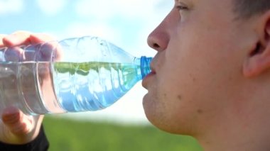 Kafkas görünümlü şişman genç bir adam şeffaf plastik bir şişeden beyaz bulutlu mavi bir gökyüzüne su içiyor. Susuzluk giderici. Şişeden temiz su iç. Yakın plan.