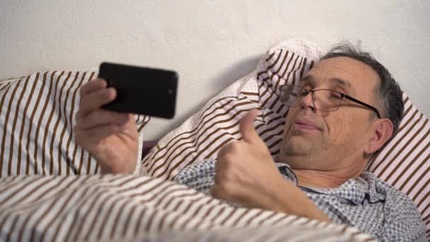 Oudere man in bed met smartphone. Communicatie op afstand met familie en vrienden via internet — Stockvideo