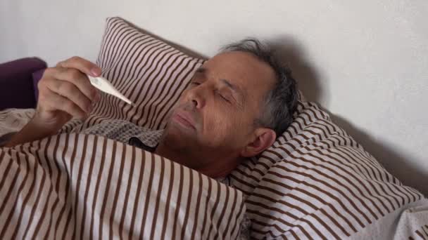 Un homme âgé regarde un thermomètre électronique alors qu'il est couché au lit. Mesurez la température de votre corps. Repos au lit — Video