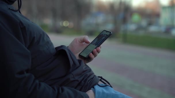 Mladý kavkazský muž používá chytrý telefon, zatímco sedí na lavičce proti městskému pozadí — Stock video