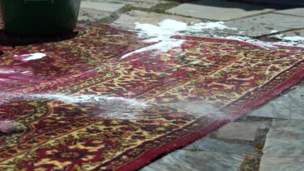 Una mujer está cepillando una alfombra sucia en la calle, en el patio de la casa de cerca. Enjuague con agua limpia. Mucha espuma del polvo. Alfombra de lavado de cerca. Cepillo limpia alfombra — Vídeos de Stock