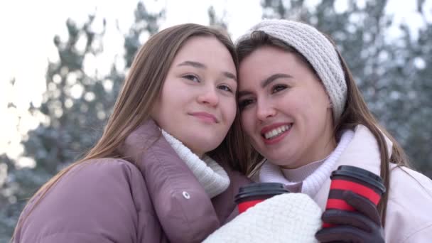 Porträt zweier glücklicher Mädchen in einem verschneiten Park an einem frostigen Wintertag. Mädchen spazieren an einem kalten, sonnigen Tag mit heißem Kaffee — Stockvideo