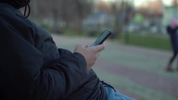 Junger Kaukasier nutzt Smartphone, während er auf Bank vor Stadthintergrund sitzt — Stockvideo