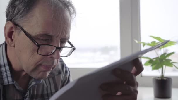 Een oudere man met een bril leest aandachtig het document op de bladen — Stockvideo