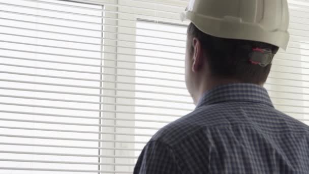 Un hombre en un casco de trabajo se encuentra junto a la ventana — Vídeos de Stock