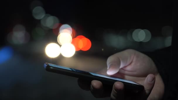 La mano de un hombre con un teléfono inteligente en la ciudad por la noche en el contexto de las luces borrosas de los coches — Vídeo de stock