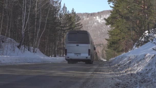 Um ônibus de passageiros passa por uma estrada coberta de neve de inverno entre montanhas arborizadas. Viaje de ônibus no inverno. Inverno dia ensolarado. — Vídeo de Stock