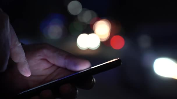 La mano de un hombre con un teléfono inteligente en la ciudad por la noche en el contexto de las luces borrosas de los coches — Vídeo de stock