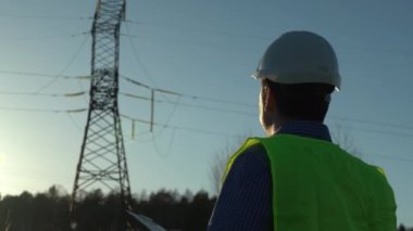 Batan güneşin ışınlarında yüksek voltajlı elektrik hatlarının yanında beyaz kasklı bir mühendis.