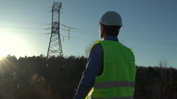 Batan güneşin ışınlarında yüksek voltajlı elektrik hatlarının yanında beyaz kasklı bir mühendis. — Stok video