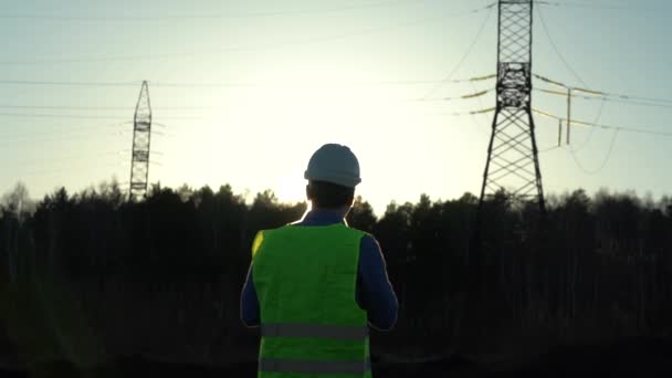 Batan güneşin ışınlarında yüksek voltajlı elektrik hatlarının yanında beyaz kasklı bir mühendis. — Stok video