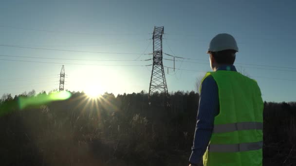 Batan güneşin ışınlarında yüksek voltajlı elektrik hatlarının yanında beyaz kasklı bir mühendis. — Stok video