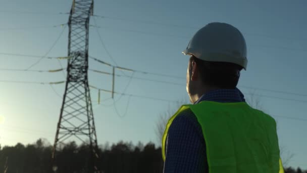 Ingénieur dans un casque blanc près des lignes électriques à haute tension dans les rayons du soleil couchant — Video