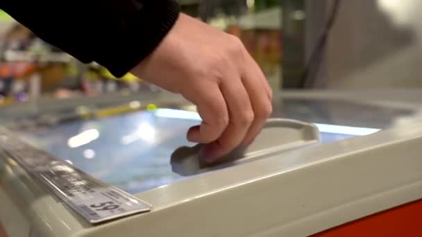 Un comprador con un carro viene y abre el refrigerador en la tienda para recuperar un paquete de salchichas del estante. El comprador selecciona comida en el refrigerador de la tienda de comestibles — Vídeos de Stock