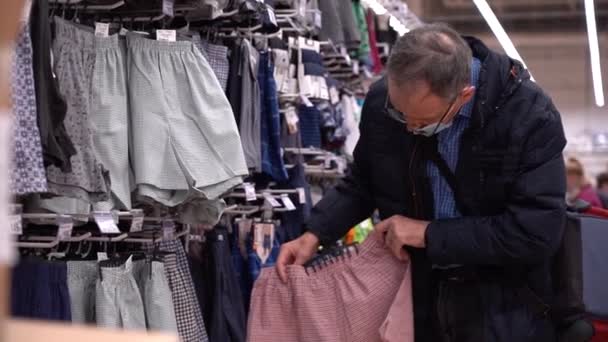 Un anciano elige ropa en una tienda. Un anciano con una máscara debido al coronavirus elige pantalones cortos para dormir en una tienda de ropa — Vídeos de Stock