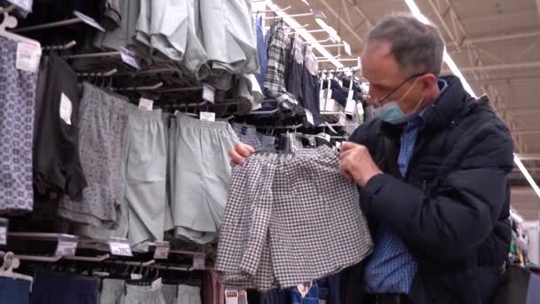 Un anciano elige ropa en una tienda. Un anciano con una máscara debido al coronavirus elige pantalones cortos para dormir en una tienda de ropa — Vídeos de Stock