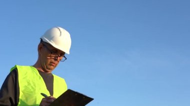 Gözlüklü ve kasklı yaşlı bir adam açık mavi gökyüzüne karşı notlar alır.