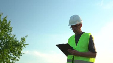 Gözlüklü ve kasklı yaşlı bir adam açık mavi gökyüzüne karşı notlar alır.