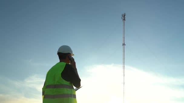 Cellular engineer in een helm met een tablet en een telefoon naast de mast. Cellulaire inspectie — Stockvideo