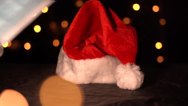 크리스마스 조명의 배경에 있는 붉은 산타클로스 캡 과 의료용 마스크 — 비디오