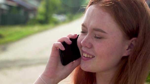 18-letá zrzavá dívka šťastně mluví po telefonu, zatímco stojí venku v letní den. Detailní záběr. — Stock video
