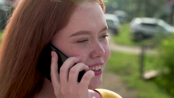 18-letnia rudowłosa dziewczyna szczęśliwie rozmawia przez telefon stojąc na zewnątrz w letni dzień. Zbliżenie. — Wideo stockowe