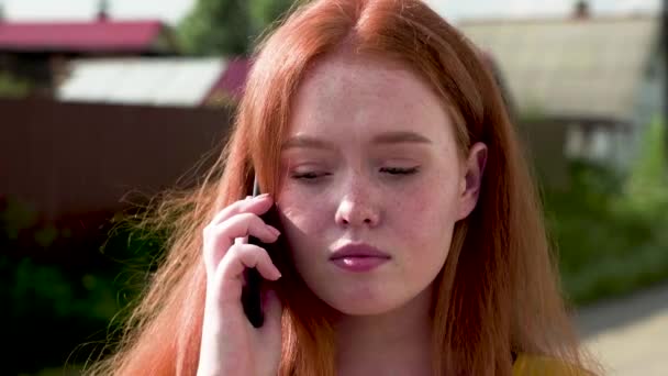 Chica de 18 años de pelo rojo con tristeza habla por teléfono, de pie en la calle en un día de verano. Primer plano — Vídeo de stock