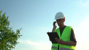 Gözlüklü ve kasklı yaşlı bir adam açık mavi gökyüzüne karşı notlar alır.