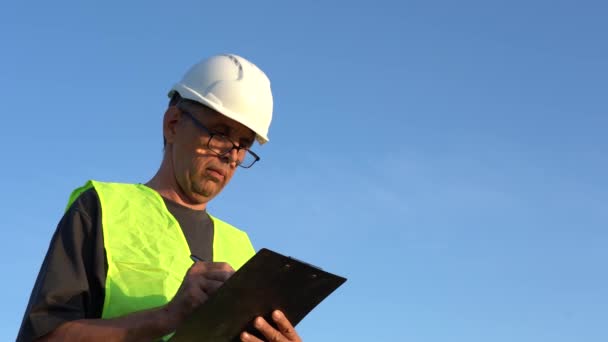 An elderly man wearing glasses and a work helmet makes notes against a clear blue sky — Stock Video