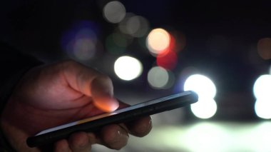Akıllı telefonu tutan ellerin yakın çekimi. Arka planda şehir ışıkları
