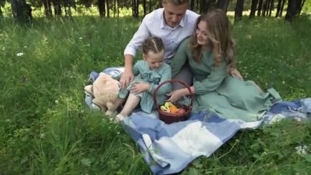 Apa, anya és kislánya egy takarón ülnek az erdő fáinak árnyékában. — Stock videók