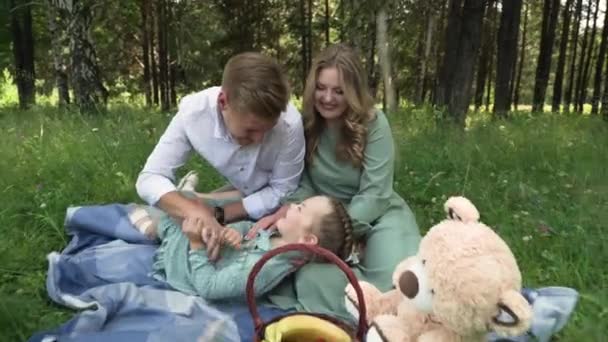 Um pai brinca com a filha. Pai, mãe e filhinha estão sentados em um cobertor à sombra das árvores na floresta — Vídeo de Stock