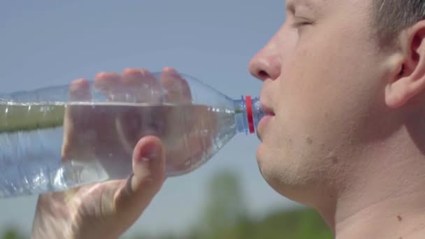 Dorst lessen. Drink schoon water uit een fles. Een jonge blanke man drinkt water uit een doorzichtige plastic fles tegen een blauwe lucht met witte wolken. Close-up — Stockvideo