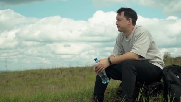 Un joven turista se sienta en una colina con una botella de agua sobre un fondo de nubes blancas. Una persona solitaria mira a la distancia y piensa en su futuro — Vídeo de stock