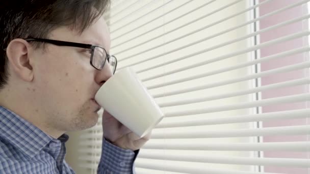 Un hombre con una taza de café se para junto a la ventana y mira a la calle. Un descanso durante el trabajo — Vídeo de stock