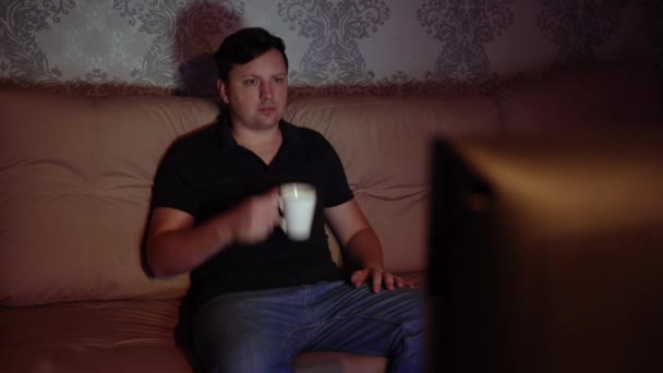 Joven hombre viendo la televisión sentado en el sofá con una taza de té está descansando por la noche — Vídeos de Stock