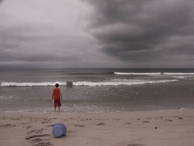 Sahilde oynayan bir çocuk. Yukarıda dramatik bulutlar var. Desenlenmiş resim.