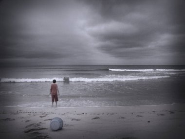 Sahilde oynayan bir çocuk. Yukarıda dramatik bulutlar var. Görüntü siyah beyaz.