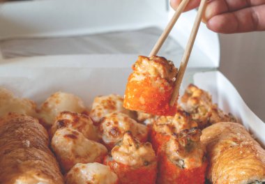 hand holding a roll with chopsticks. Lunch boxes, baked sushi set. Assorted rolls on a light background. Restaurant delivery, takeaway concept. Japanese seafood cuisine