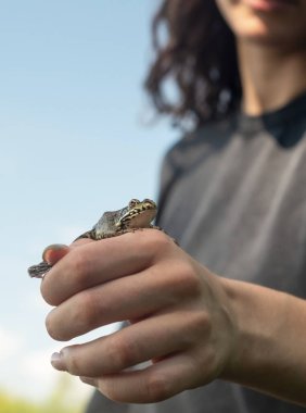 Bir kız elinde küçük vahşi bir kurbağa tutuyor gölün arka planında.