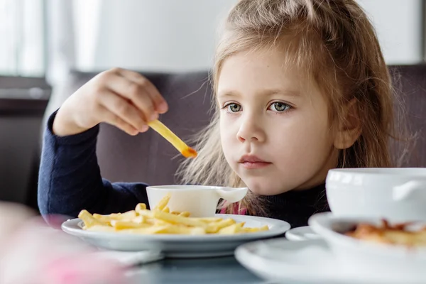 Liten flicka sitter vid bord och äter pommes frites — Stockfoto