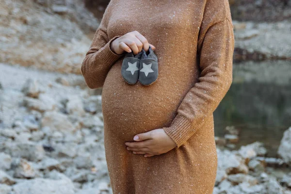 Kahverengi kazaklı hamile bir kadın, elinde botları, vücudunun parçaları, plajın arka planında, bir çocuğu bekleme, ebeveynler, ebeveynlik ve yeni doğmuş bir bebeğe bakma kavramı.