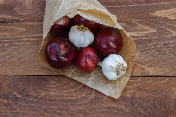Sebzeler kağıt torbada kırmızı soğan ve sarımsak. Kahverengi ahşap arka planda yakın plan. Metin, pişirme konsepti, manav, vitaminler, sağlık ve bağışıklık için boş alan var.