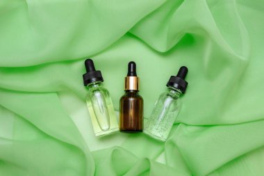 Yağ, serum ve yeşil ipek kumaşlı pipeti olan üç kozmetik cam şişe. Kapalı, düz, düz, yüz ve vücut bakımı konsepti, kadınların yaşlanmayı önleyici kozmetik ürünleri, kopyalama alanı.