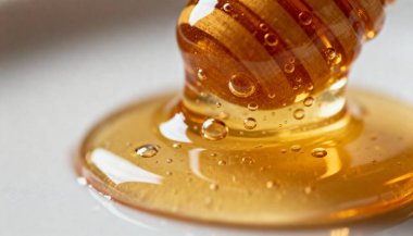 Tahta bir baldan damlayan altın balın yüksek çözünürlüklü makro fotoğrafçılığı beyaz bir yüzey üzerinde parlak, tatlı bir sıvı havuzu oluşturur. Organik yiyecekler, sağlıklı doğal tatlılık..